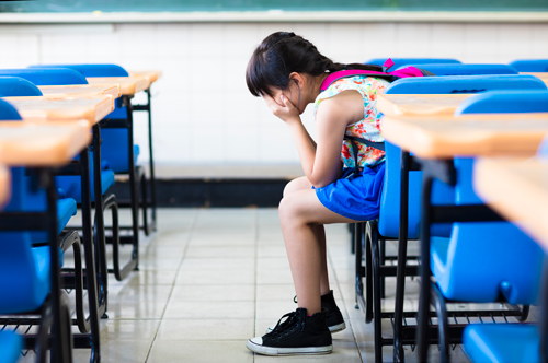 A young student sits alone in a classroom with her head in her hands.